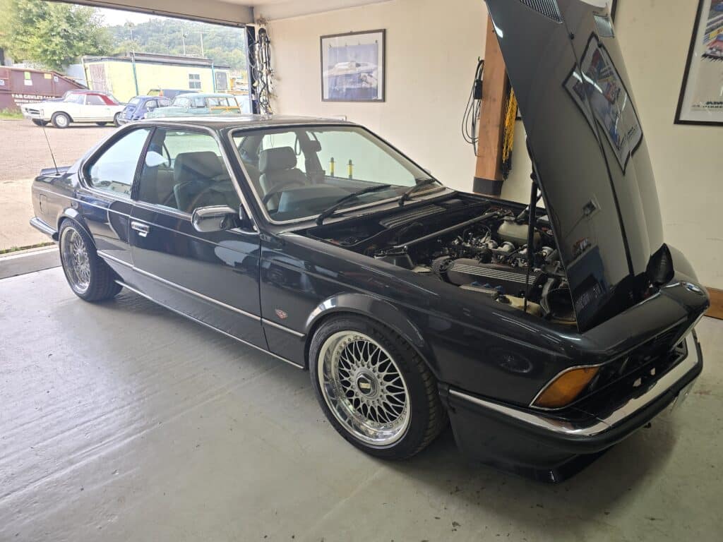 A classic BMW 6 Series coupe in glossy black, parked inside ASD Tuning's workshop with its bonnet open, revealing a modified engine. The car features BBS-style multi-spoke wheels, a lowered stance, and a well-maintained body. The vehicle is running an ECUMaster Classic ECU.