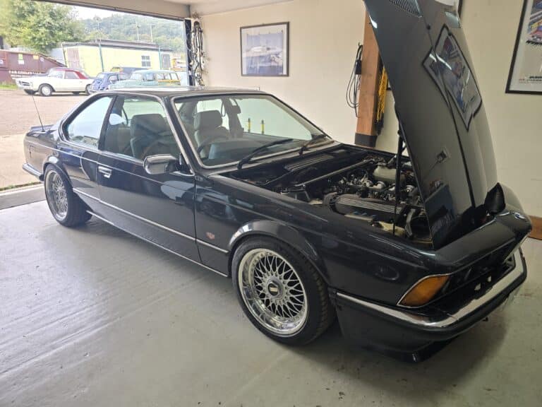 A classic BMW 6 Series coupe in glossy black, parked inside ASD Tuning's workshop with its bonnet open, revealing a modified engine. The car features BBS-style multi-spoke wheels, a lowered stance, and a well-maintained body. The vehicle is running an ECUMaster Classic ECU.